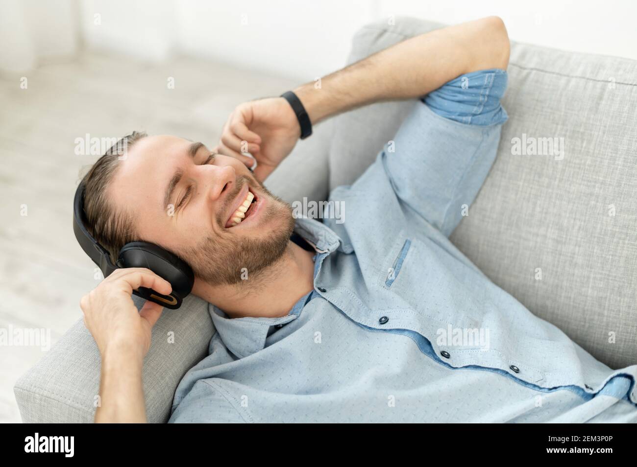 Homme attrayant avec des cheveux et une barbe marron clair, chantant sa  chanson préférée, souriant, écoutant de la musique avec des écouteurs  couchés sur le canapé gris confortable Photo Stock - Alamy