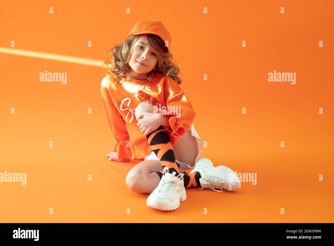 Fille de 6 ans, assise sur le sol en studio sur fond orange, posant dans des vêtements décontractés, une casquette et des baskets. Enfance, mode, publicité, sp Banque D'Images