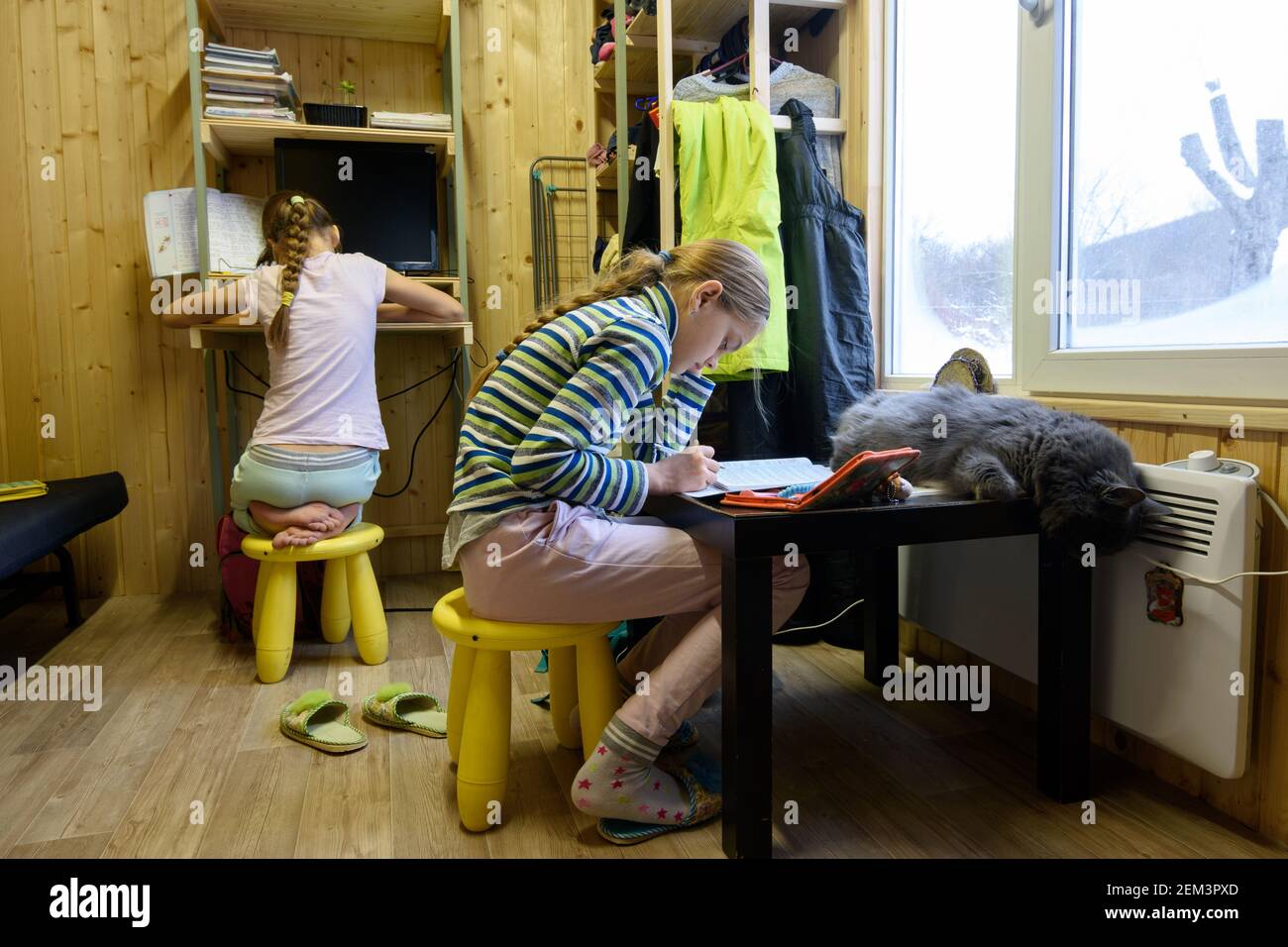 Deux filles font des devoirs à la table d'ordinateur et la table sur laquelle le chat dort par la fenêtre dans une petite pièce d'une maison de campagne Banque D'Images