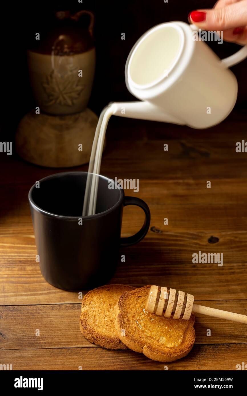 Une main femelle verse du lait d'un pot dans un tasse avec des roussins et du miel à côté Banque D'Images