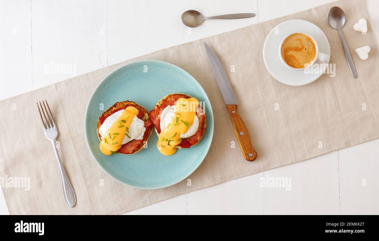 Œufs Benedict, muffins anglais, jambon grillé, œufs pochés, sauce hollandaise, herbes de ciboulette, citron, tasse de café. Table en bois blanc avec petit déjeuner. Bas Banque D'Images