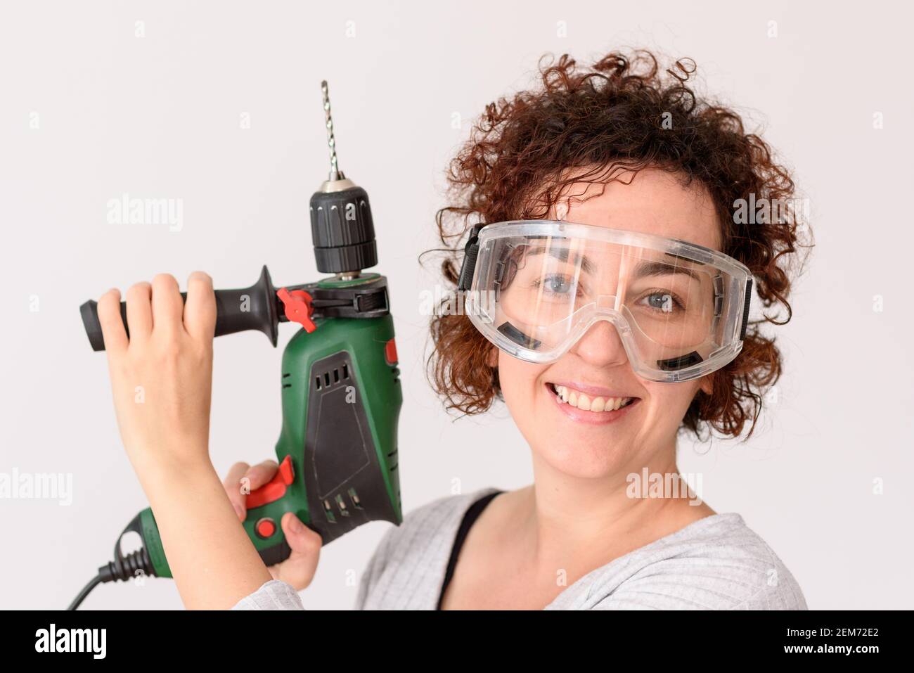 Une femme caucasienne aux cheveux bouclés tient un exercice dans ses mains, prête à faire des améliorations à la maison. Elle est debout devant un établi. Elle porte un gris Banque D'Images