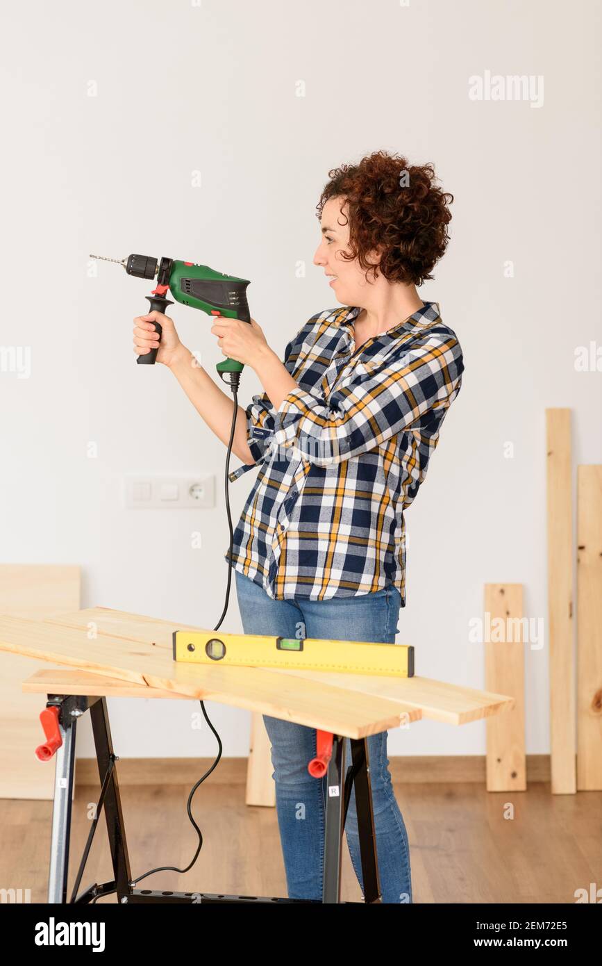 Une femme caucasienne aux cheveux bouclés tient un exercice dans ses mains, prête à faire des améliorations à la maison. Elle est debout devant un établi. Elle porte un plai Banque D'Images