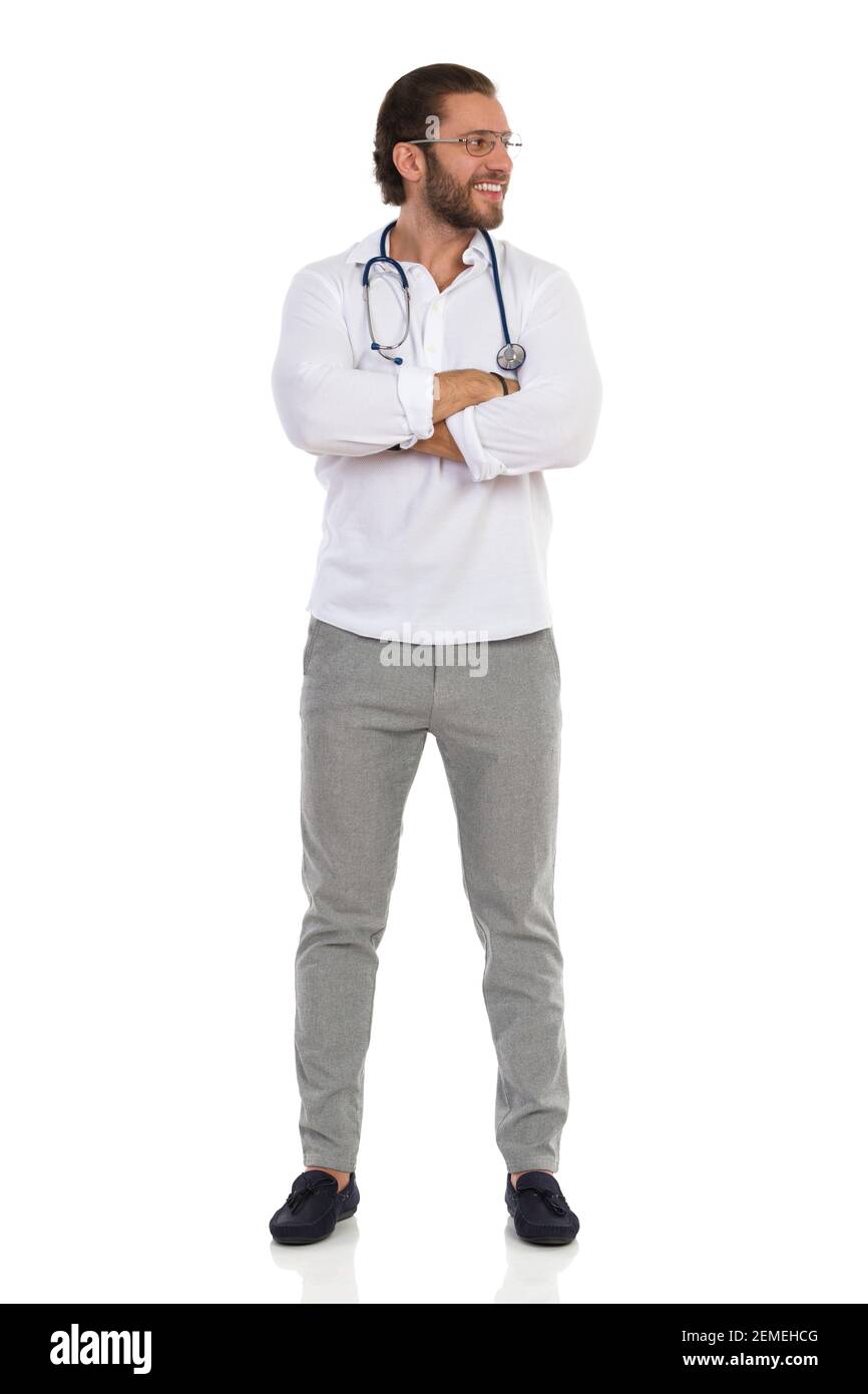 Un jeune médecin souriant est debout avec les bras croisés, regardant sur le côté. Vue avant. Prise de vue en studio sur toute la longueur isolée sur blanc. Banque D'Images