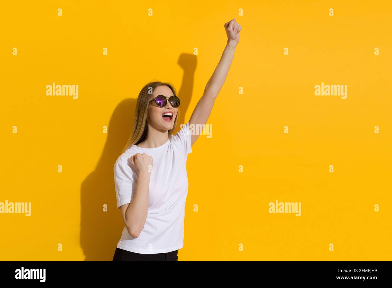 Bonne jeune femme blonde en chemise blanche et lunettes de soleil est debout avec le poing élevé et criant. Taille relevée sur fond jaune. Banque D'Images