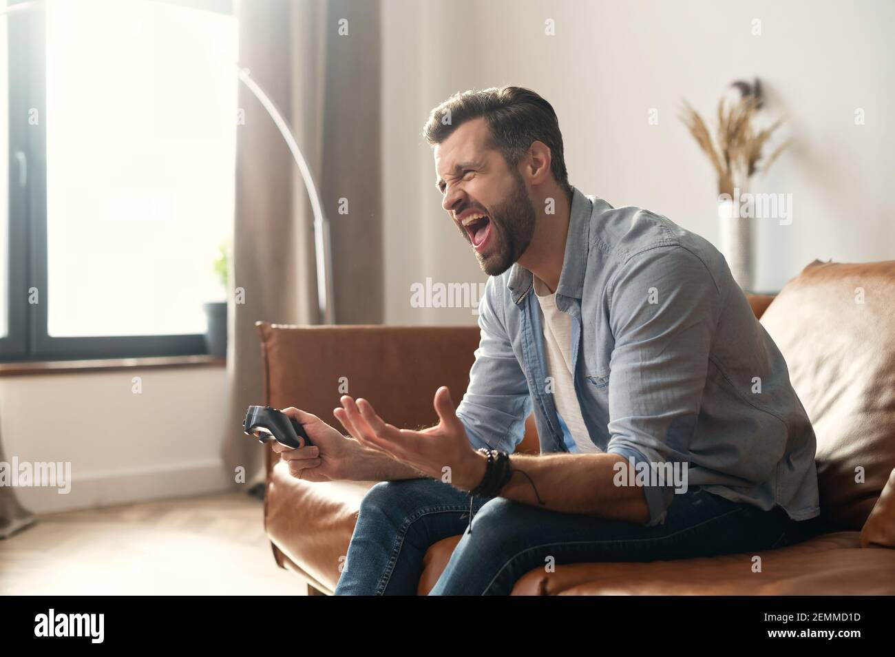 Vue latérale portrait d'un jeune homme hippster barbu assis sur le canapé dans le salon, tenant le joystick, hurlant en raison de la partie perdue de jeu vidéo, le gamer accro mâle se sentant fou, gesturant Banque D'Images