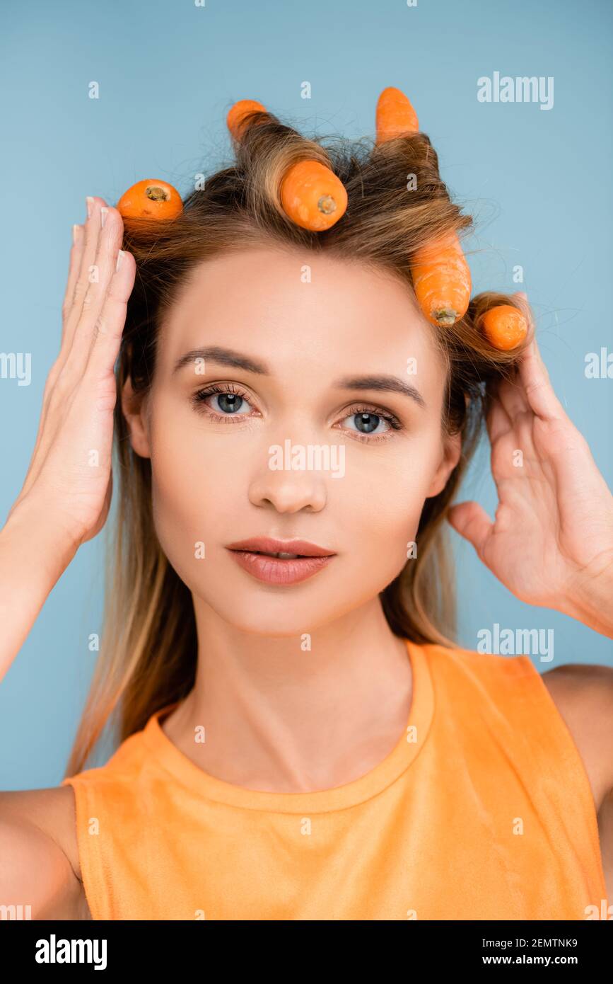 jolie femme au maquillage naturel utilisant des carottes comme des curlers isolés sur bleu Banque D'Images