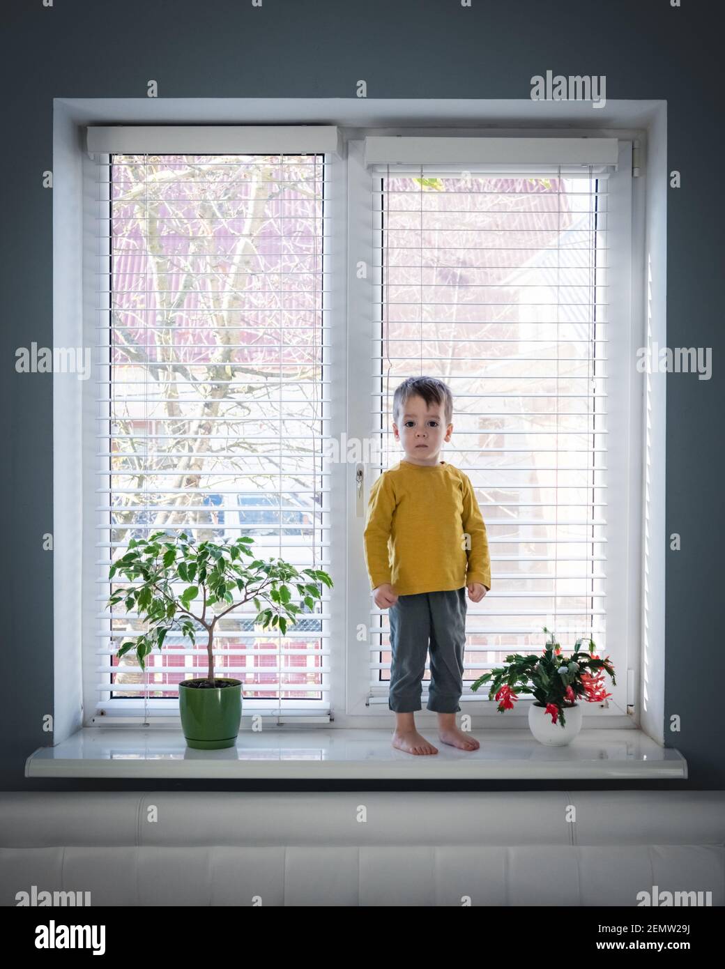 Un petit garçon solitaire reste sur le rebord de la fenêtre près d'une grande fenêtre dans sa chambre. Concept de l'enfance Banque D'Images