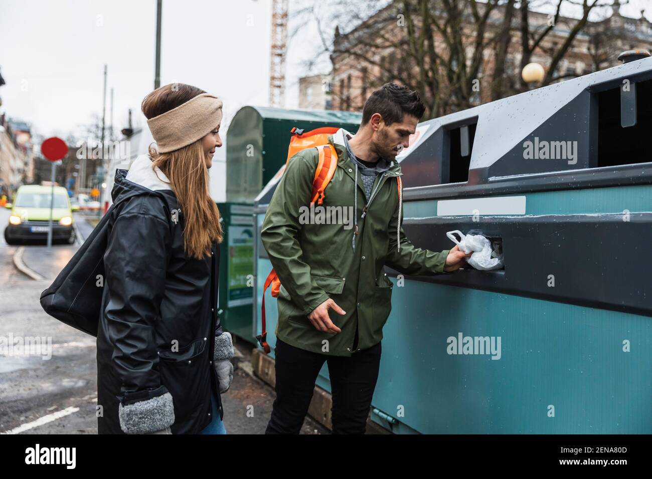 Couple hétérosexuel jetant du plastique dans un bac de recyclage tout en se tenant à ville Banque D'Images