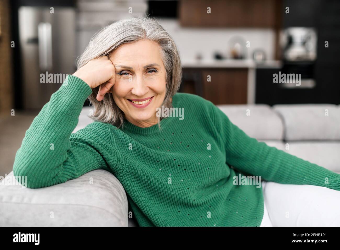 Femme âgée souriante avec des cheveux gris bien entretenus, des yeux bleus et de belles rides, assise sur le canapé à la maison, tête reposant sur une main et regardant l'appareil photo, se sent calme et équilibré, amical Banque D'Images