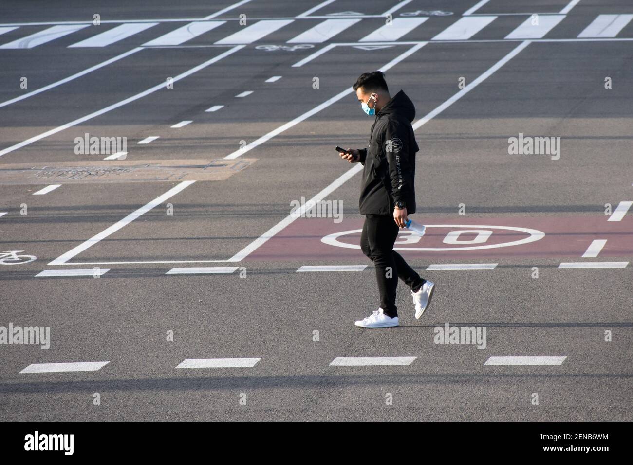 Jeune homme avec masque traversant une rue pendant qu'il vérifie son smartphone. Banque D'Images