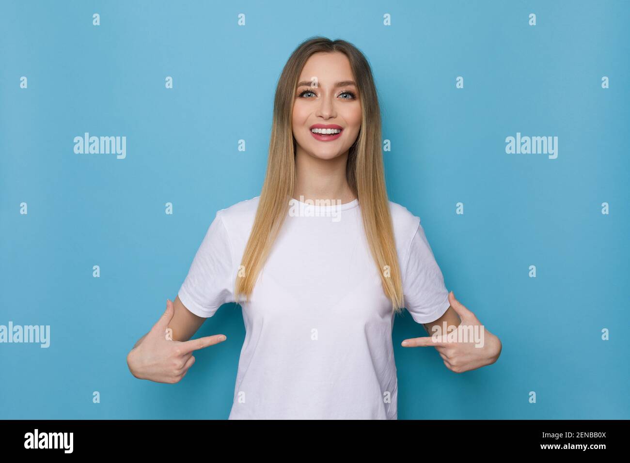 Une fille souriante en chemise blanche se pointe vers elle-même. Taille haute studio tourné sur fond bleu. Banque D'Images