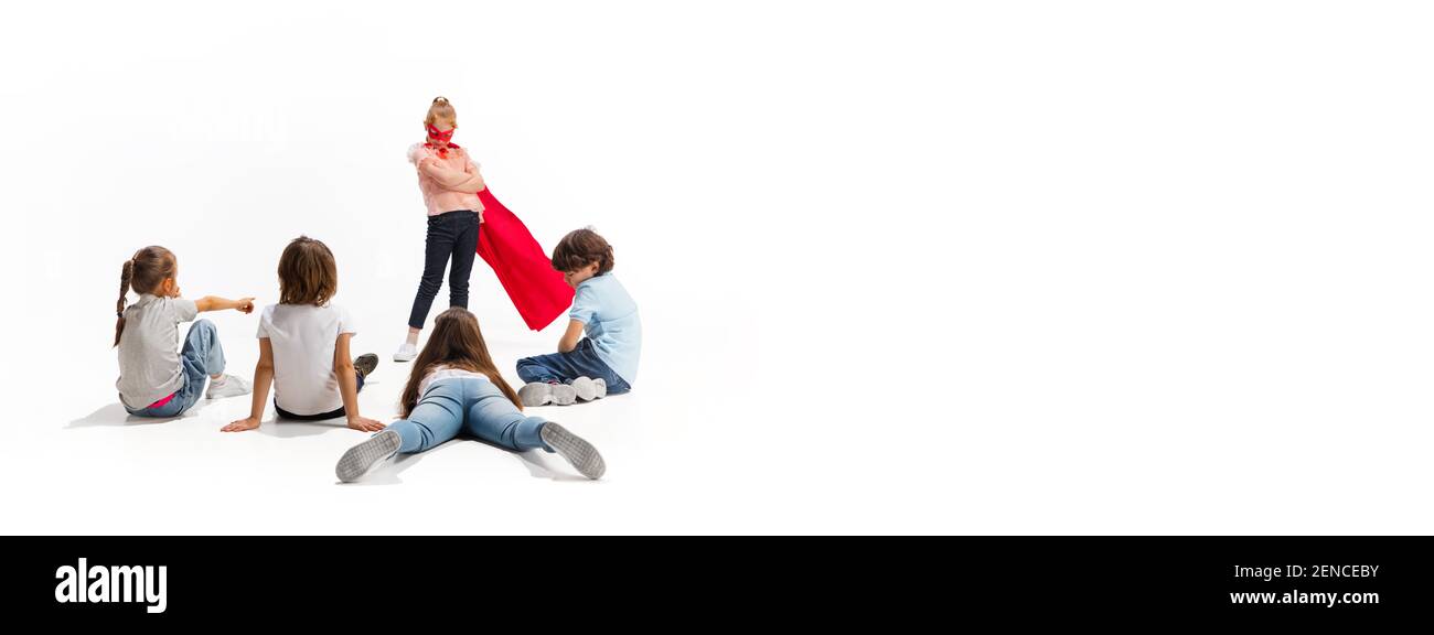 Circulaire. Enfant prétendant être un super-héros avec ses amis assis autour d'elle. Les enfants sont excités, inspirés par leur ami robuste et courageux, sous un manteau rouge isolé sur fond blanc. Rêves, émotions concept. Banque D'Images