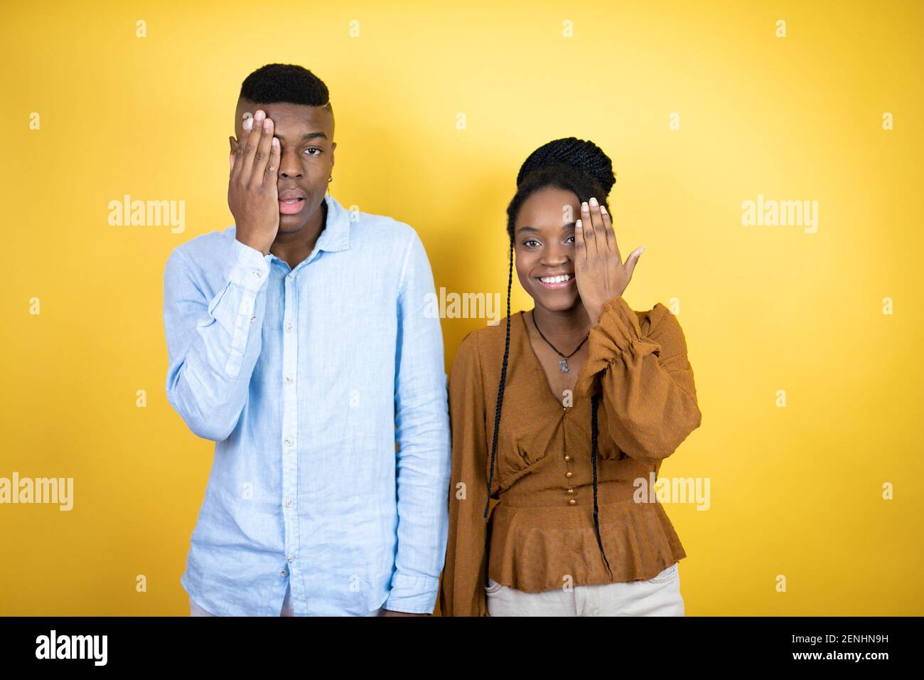 Jeune couple afro-américain debout sur fond jaune couvrant un oeil avec la main, sourire confiant sur le visage et émotion surprise. Banque D'Images