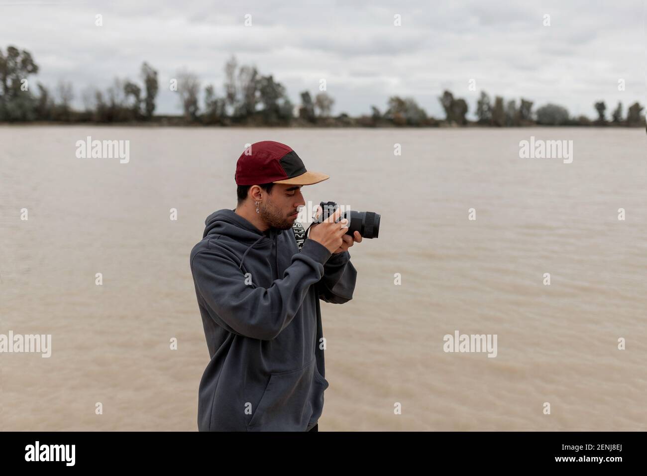 Jeune homme portant un chandail gris et une casquette rouge est prendre des photos sur un quai à côté d'une rivière Banque D'Images