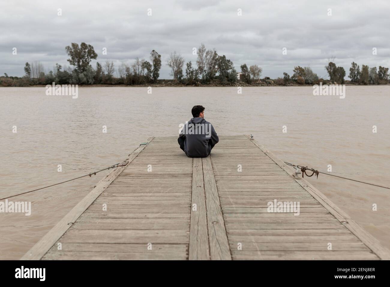 Jeune homme de derrière assis dans un quai à côté de une rivière tout en regardant et en se reposant avec un ciel nuageux Banque D'Images