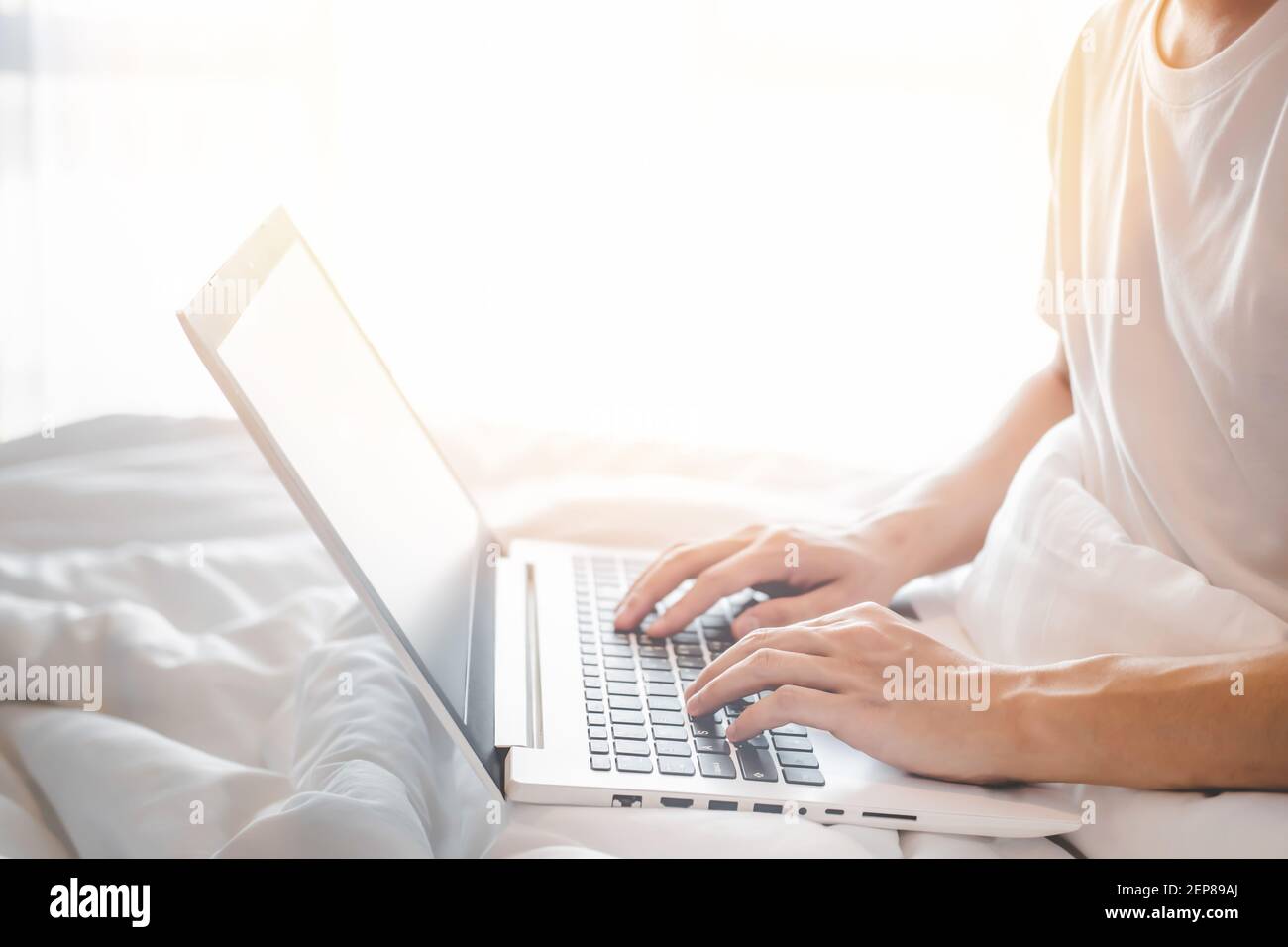 Gros plan d'un jeune homme portant un t-shirt blanc à manches courtes à l'aide d'un ordinateur portable et assis sur un lit blanc et une couverture blanche avec lumière du soleil. Concept de travail de h Banque D'Images