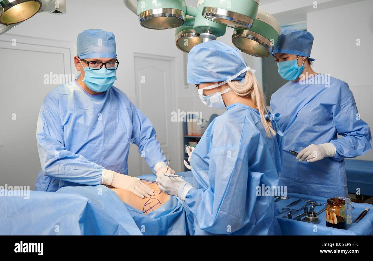 Groupe de médecins en uniforme chirurgical bleu effectuant la chirurgie cosmétique dans la salle d'opération. Équipe chirurgicale effectuant une chirurgie esthétique et utilisant des instruments médicaux en clinique. Concept de chirurgie plastique. Banque D'Images