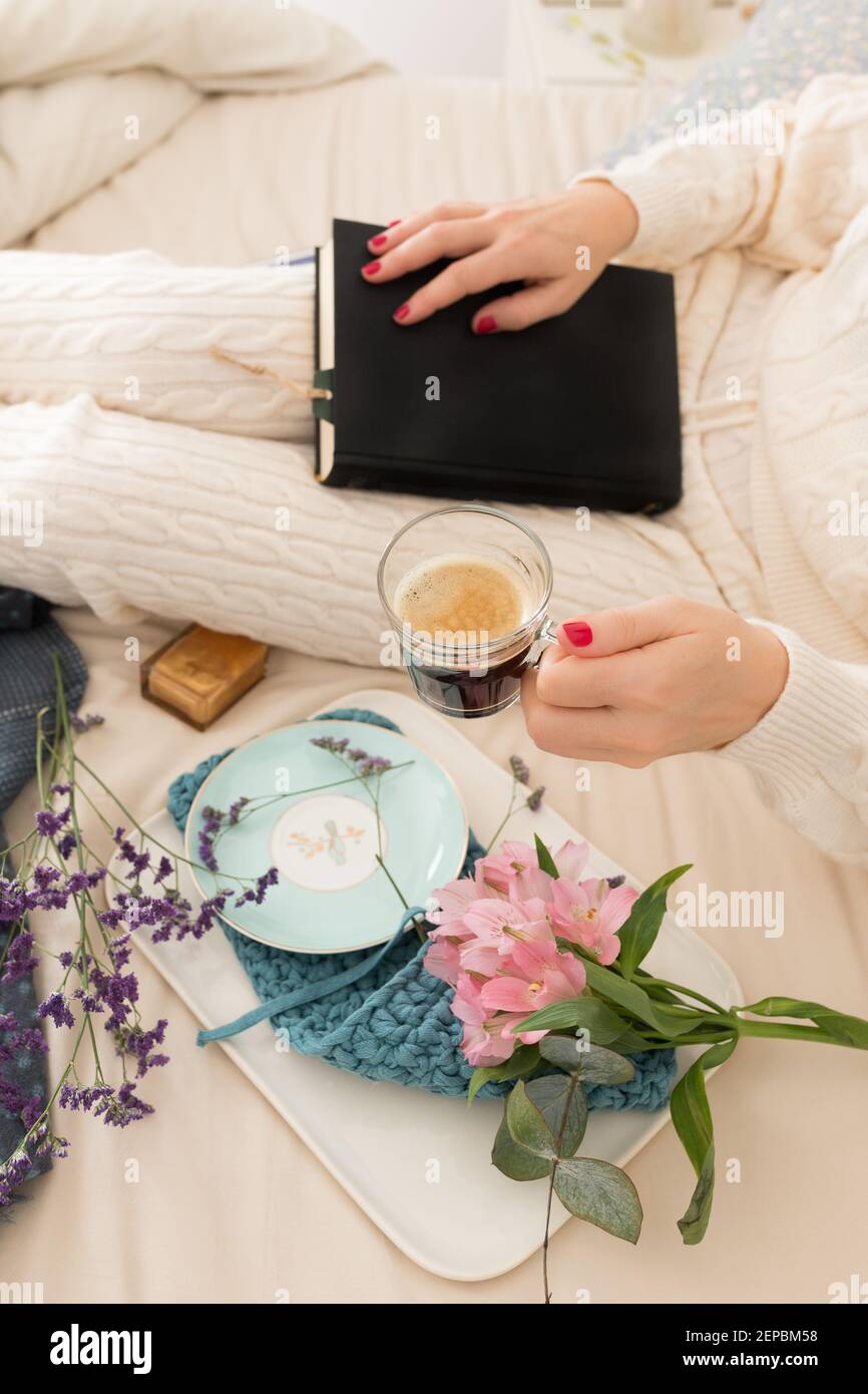 Femme vêtue de vêtements de détente chauds sur le lit avec un livre fermé sur ses genoux. Elle tient une tasse de café mousseux. Banque D'Images