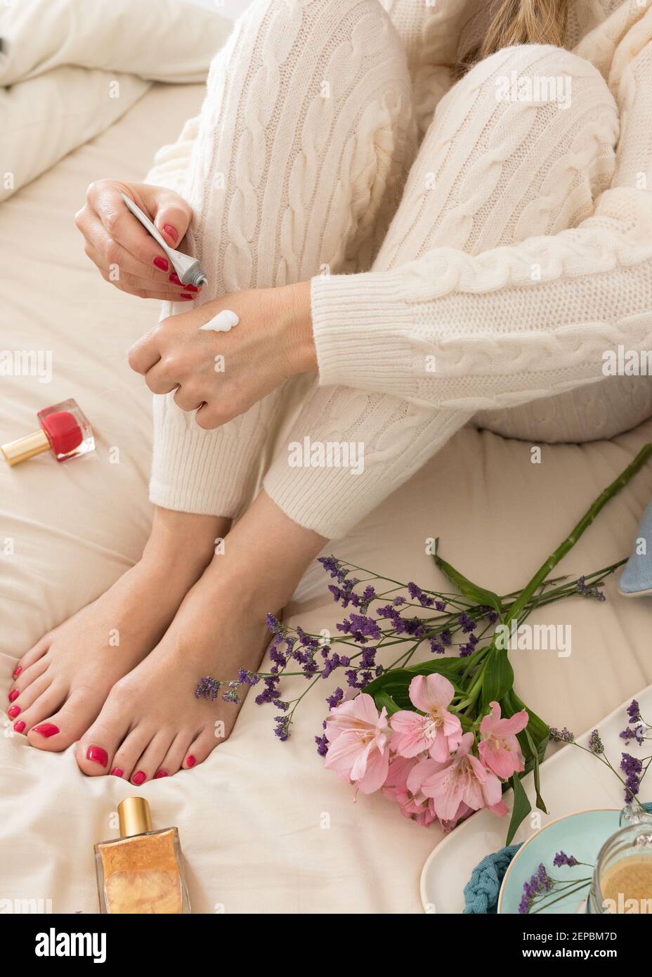 Un tube de crème main tenu par une main peinte rouge ongles main d'une femme vêtue de laine chaude de salon appliquant de la crème dans l'autre main. Banque D'Images