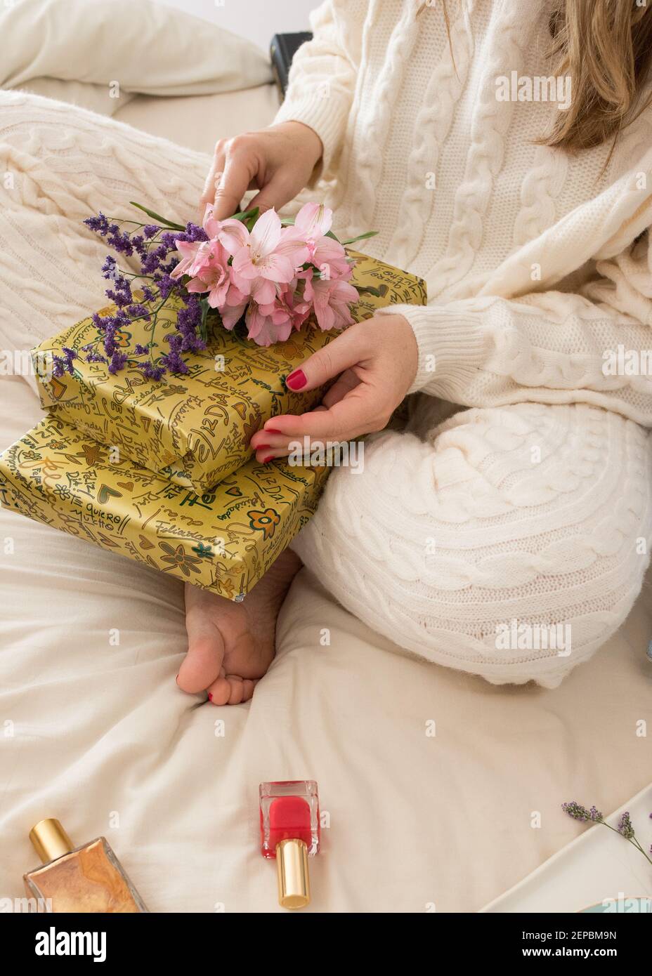 Une femme vêtue de confortables vêtements de détente en laine a deux cadeaux et un bouquet de haut fleuri sur ses genoux. Banque D'Images