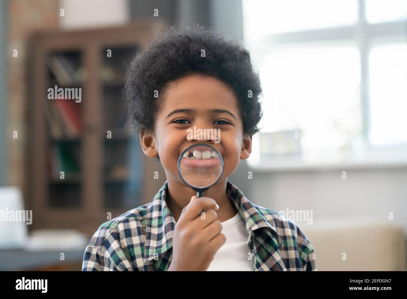 Adorable écolier d'origine africaine tenant une loupe devant de sa bouche et de montrer son sourire en regardant à vous Banque D'Images