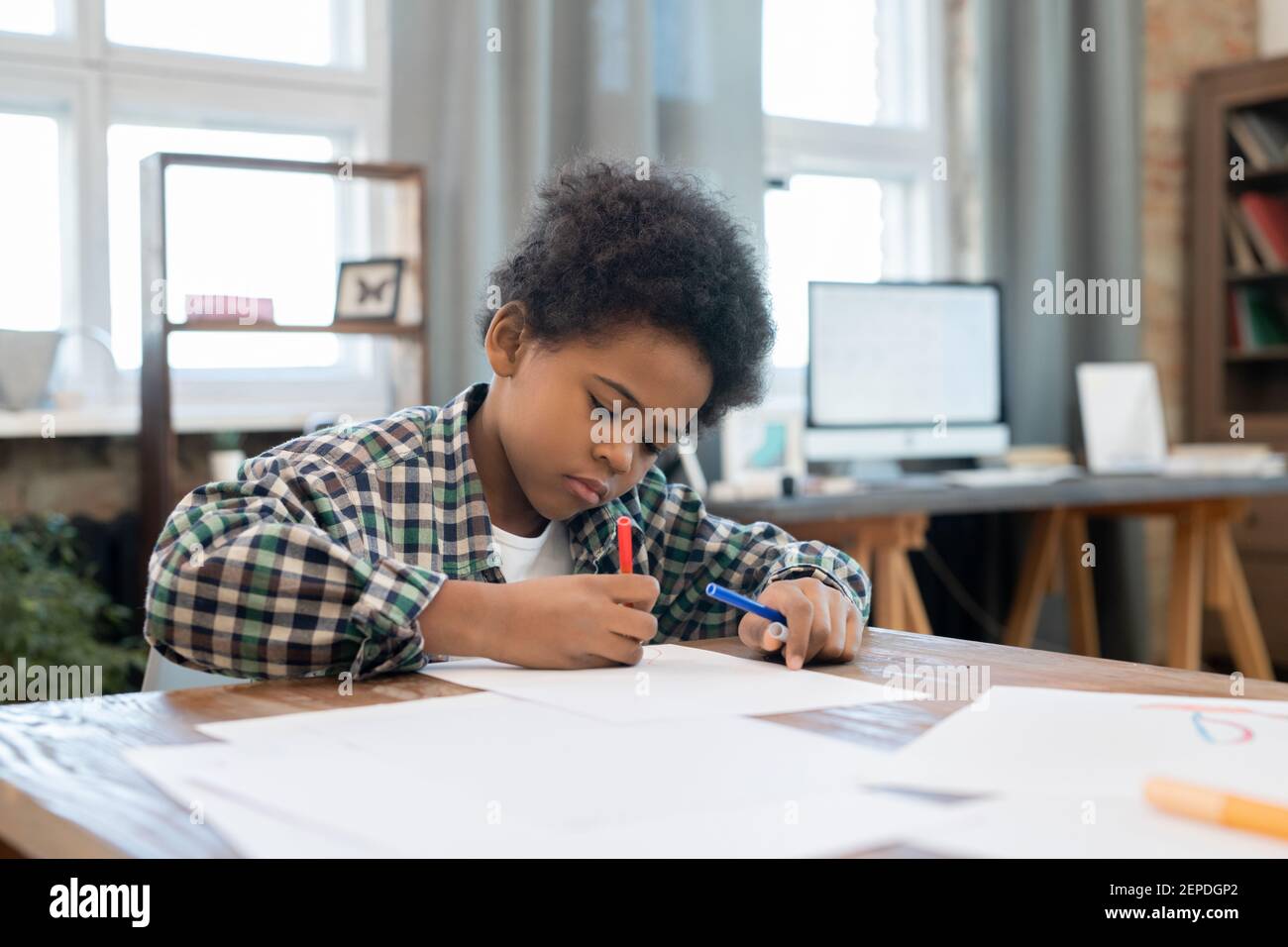 Jeune garçon diligent en vêtements décontractés, dessin avec surligneurs sur papier par table dans le salon tout en appréciant les loisirs après avoir fait vos devoirs Banque D'Images