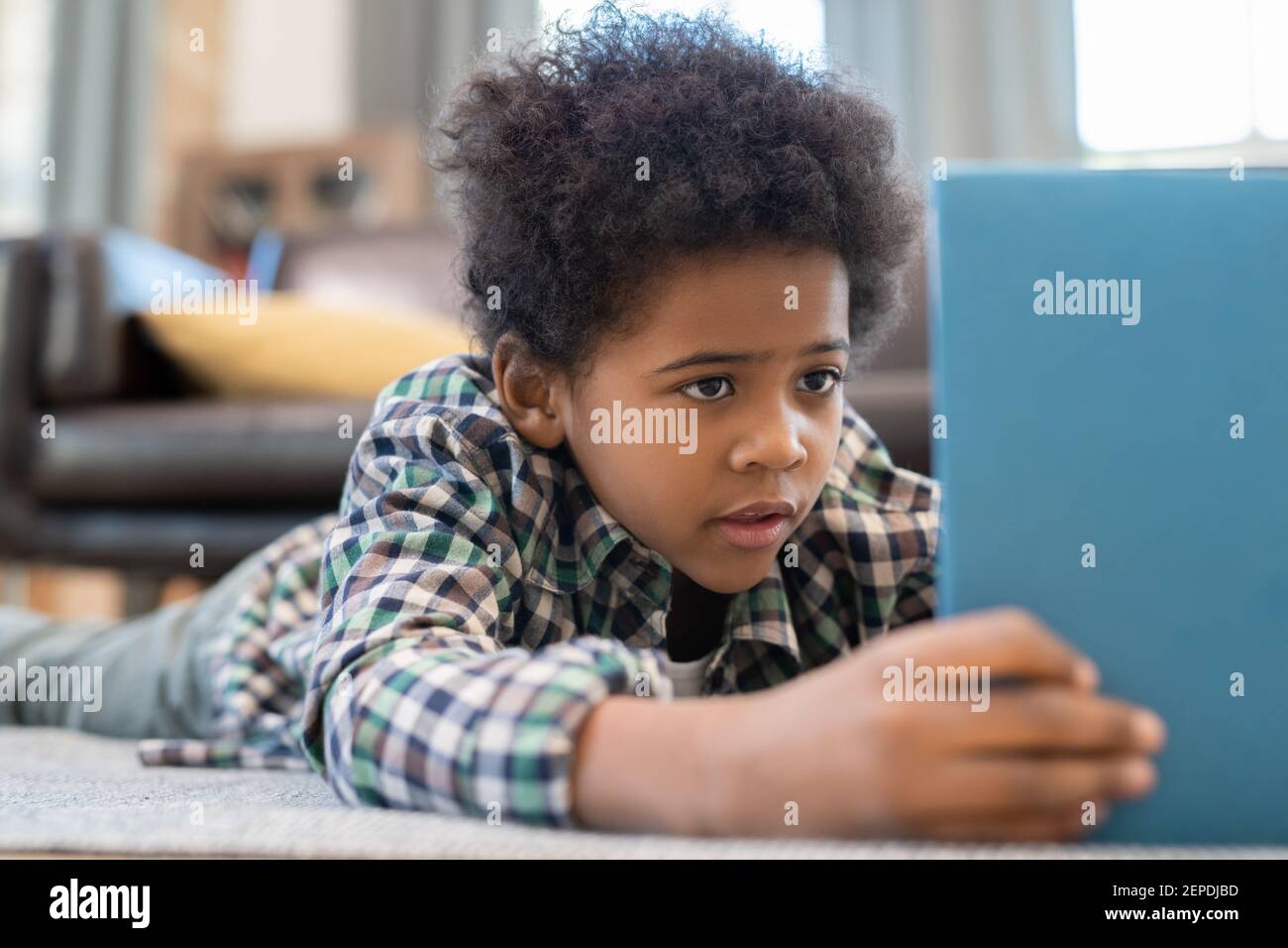 Un adorable petit garçon d'école élémentaire avec une tablette numérique posée sur le sol contre le canapé en cuir noir dans le salon et regarder curieux en ligne vidéo Banque D'Images