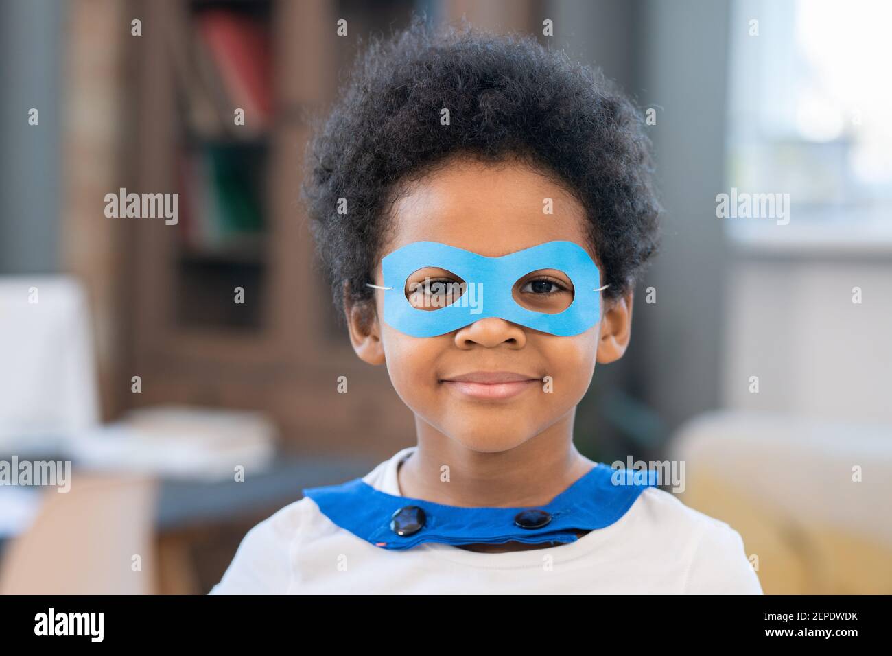 Mignon garçon de course mixte en costume de super-héros qui vous regarde à travers les lunettes de papier bleu drôle tout en se tenant devant appareil photo Banque D'Images