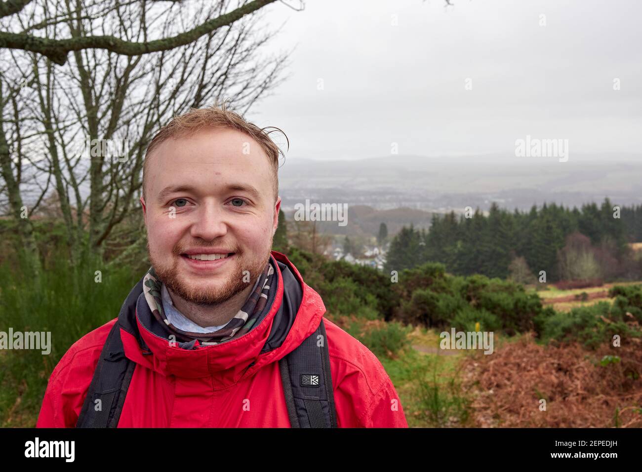 Un randonneur masculin sous la pluie souriant dans une veste rouge, avec un sac à dos, une barbe en face d'une vallée dans des conditions météorologiques défavorables dans les Highlands. Banque D'Images