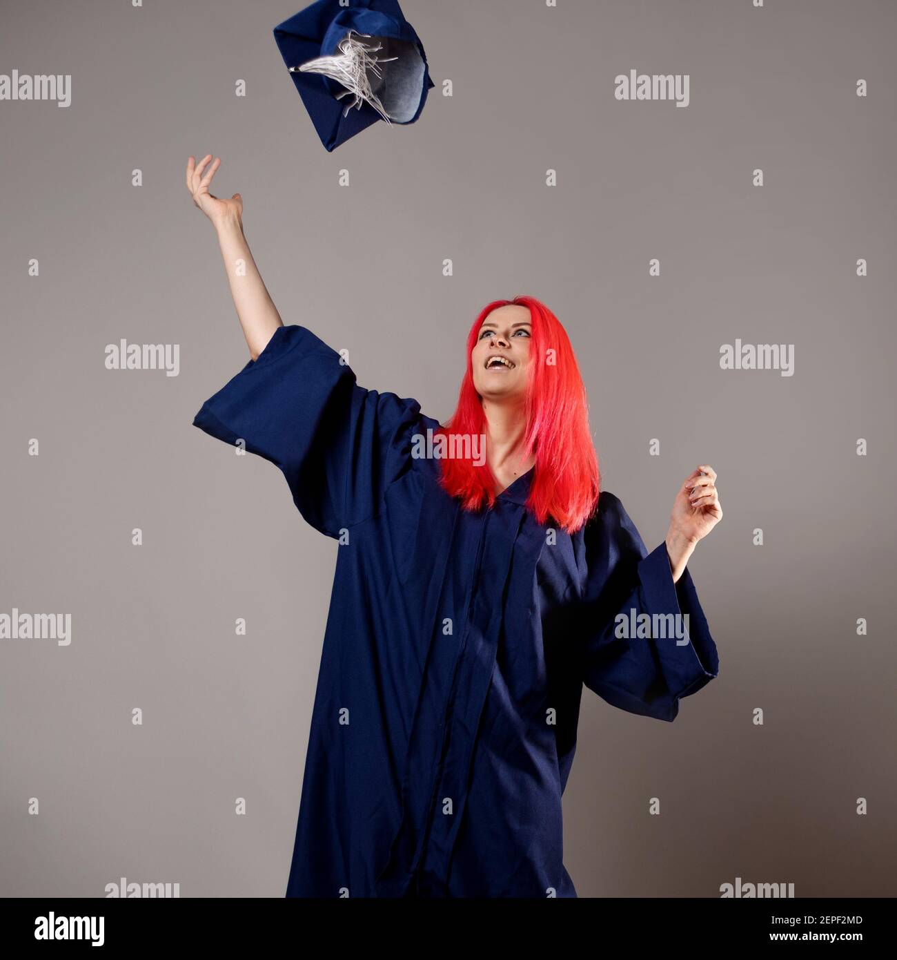 Bon diplômé sur fond gris. Une jeune femme aux cheveux brillants avec une casquette et une robe. Jetez le capuchon. Banque D'Images
