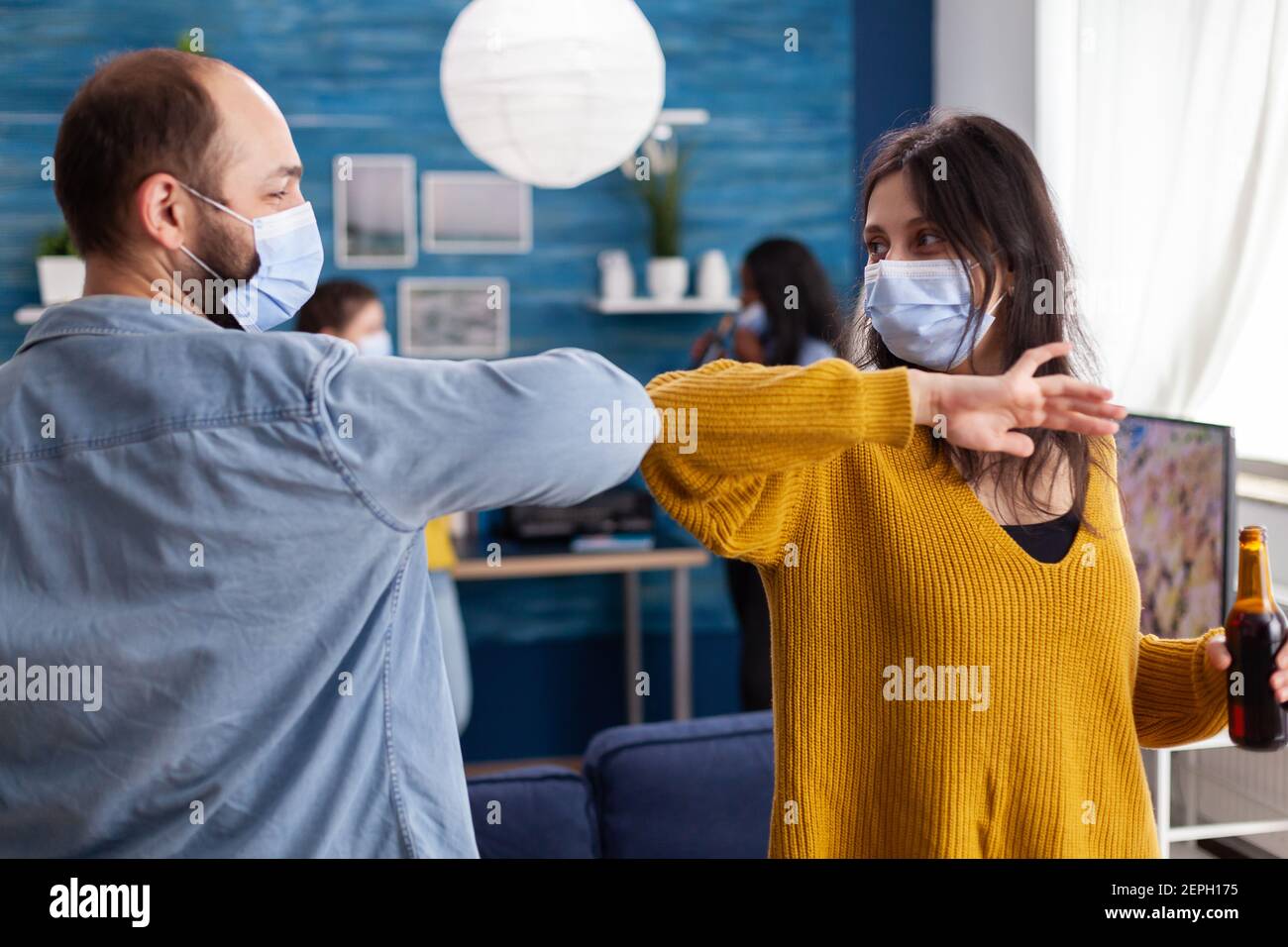 Heureux amis gaies touchant le coude en gardant social distancer salutation eachautre port masque facial empêchant la propagation du coronavirus tenant des bouteilles de bière dans le salon de l'appartement. Image conceptuelle. Banque D'Images