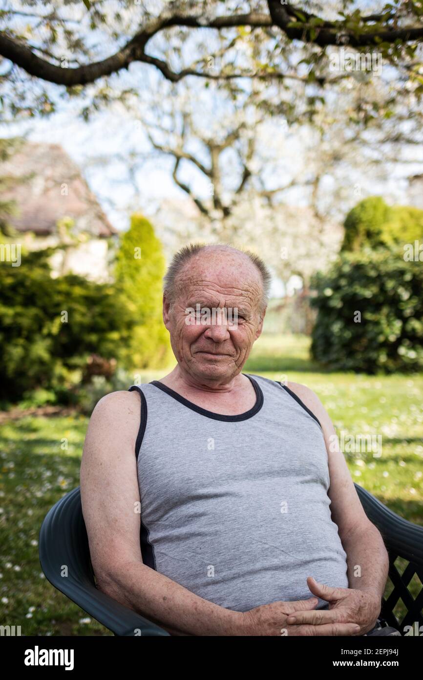 Portrait d'un homme âgé heureux assis sur une chaise dans son jardin. Vieil homme avec visage souriant se détendre dans l'arrière-cour au printemps. Regarder l'appareil photo Banque D'Images