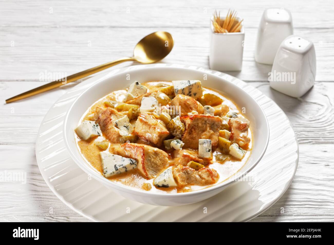 Cuisine allemande : ragoût de porc à la crème, cornichons et fromage bleu, paprika fumé servi sur une assiette blanche avec une cuillère dorée sur un dos en bois blanc Banque D'Images