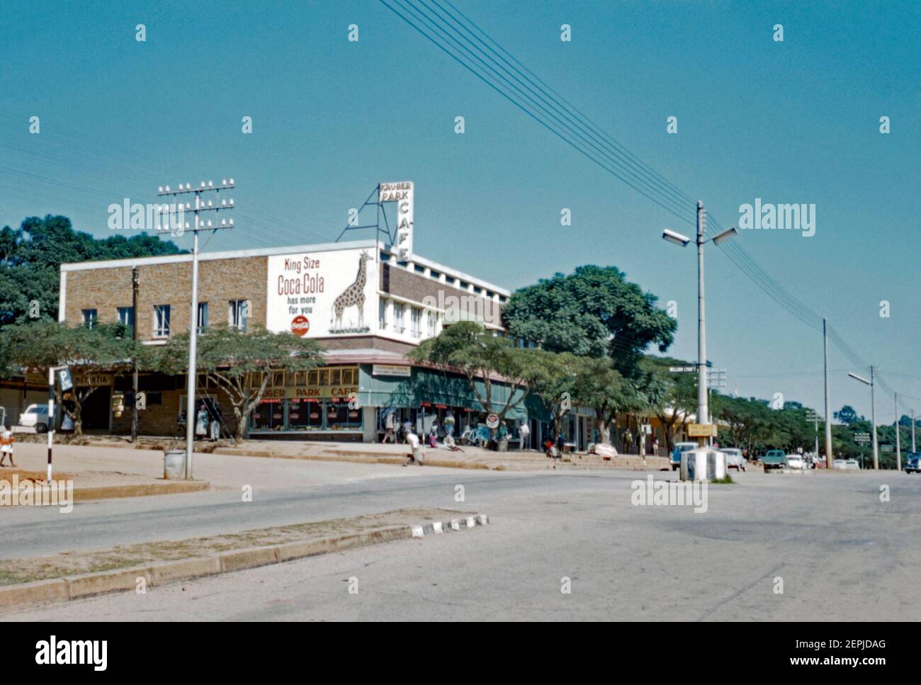 Vue sur la rue Tom Lawrence, White River (Witrivier), province de Mpumalanga, Afrique du Sud en 1962. La ville est une petite ville de vacances et d'agriculture située juste au nord de Mbombela dans l'ancienne province de Transvaal. Les fermes de la région produisent des fruits tropicaux, des légumes, des fleurs et du bois. Le bâtiment situé à l'angle de la rue William Lynn indique que le Kruger Park café y était situé. Un grand panneau d'affichage Coca Cola présente une girafe. Cette image provient d'une transparence de couleur 35 mm amateur ancienne. Banque D'Images