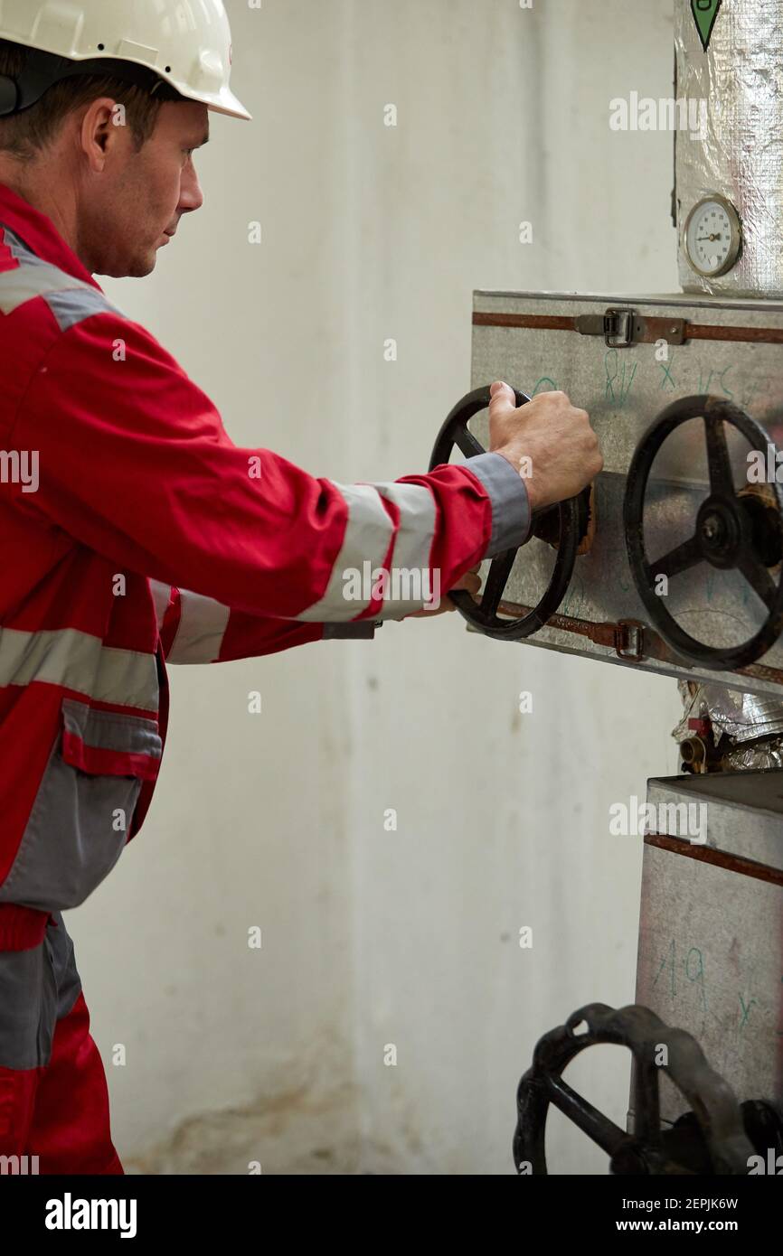 Industrie de l'énergie. Un technicien vêtu de combinaisons rouges et un casque blanc vérifiant les paramètres de chauffage. Technologie de distribution de l'eau de chauffage. Banque D'Images