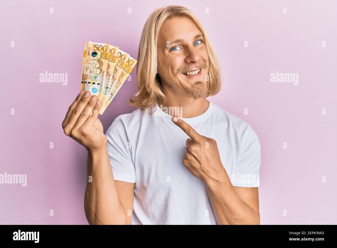 Jeune homme caucasien avec de longs cheveux tenant 500 peso philippin les billets souriants sont souriants et se mettent à pointer avec la main et le doigt Banque D'Images