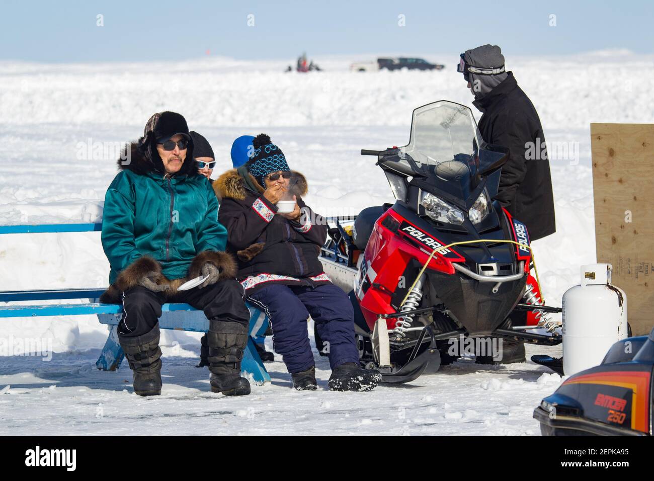 Autochtones assis sur un banc à l'extérieur près d'une machine à neige, festival d'hiver de Beluga Jamboree, Tuktoyaktuk, Territoires du Nord-Ouest, Arctique canadien. Banque D'Images