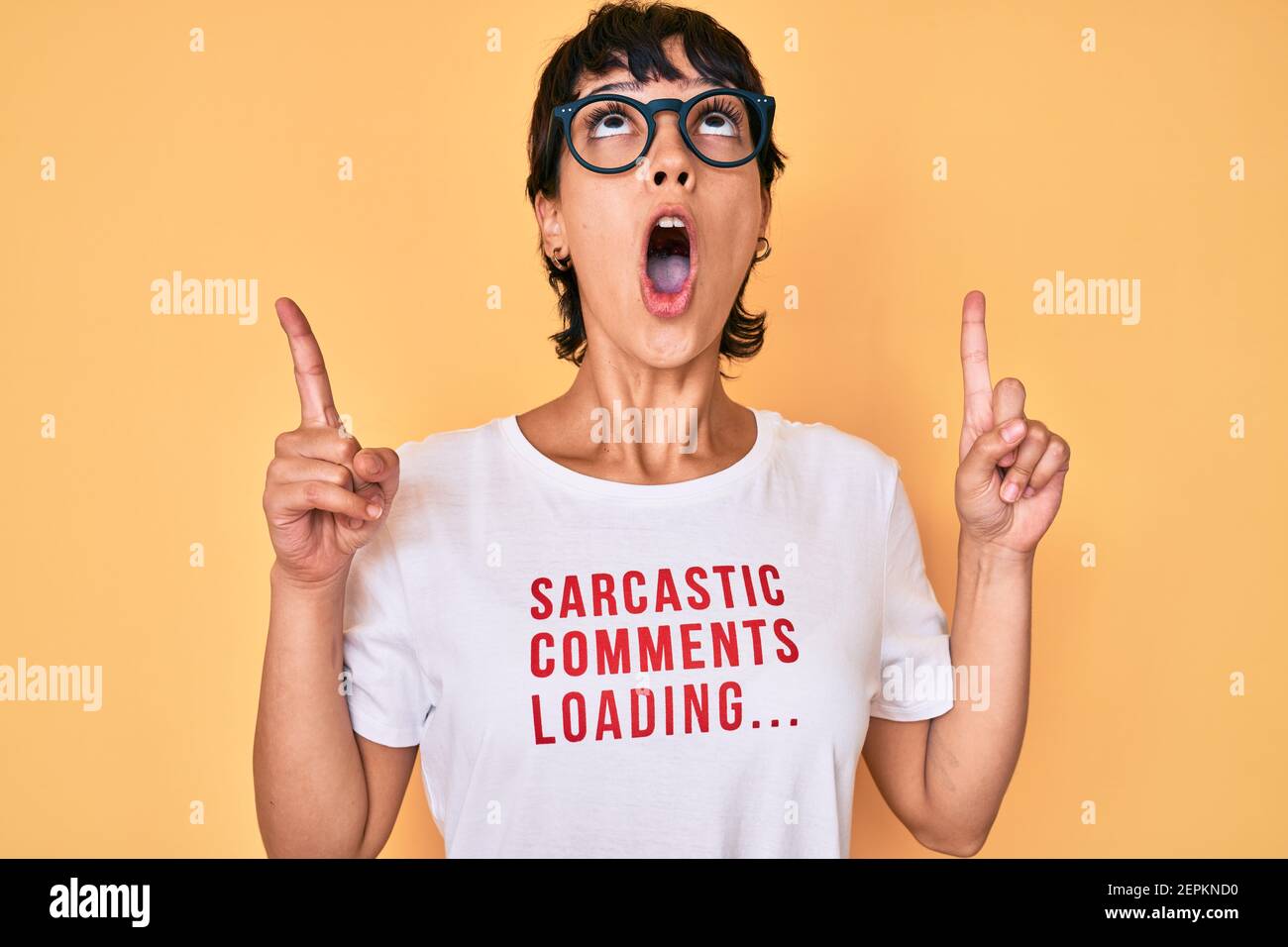 Belle femme brunettte portant des commentaires sarcastiques chargement du t-shirt stupéfait et surpris regardant vers le haut et pointant avec les doigts et les bras relevés. Banque D'Images