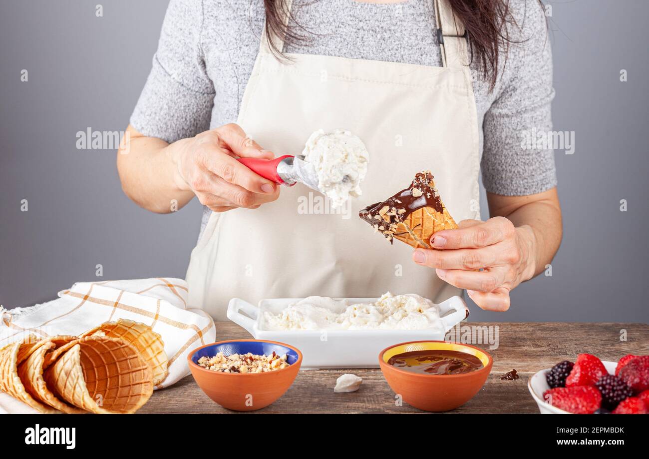 Une femme chef ramasse de glace à la vanille maison sur des cornets de glace à gaufres faits main. Elle a plongé les cônes dans le chocolat fondu et les morceaux de noix f Banque D'Images