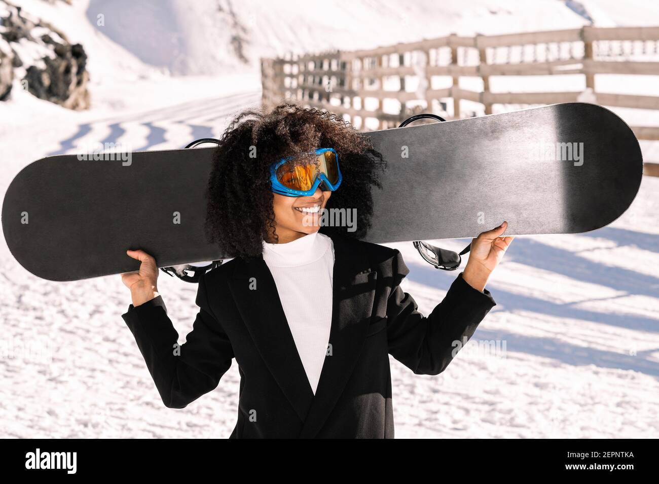 Happy Ethnic Sportswoman en vêtements élégants et lunettes de protection pour le transport du snowboard sur un support enneigé au soleil Banque D'Images