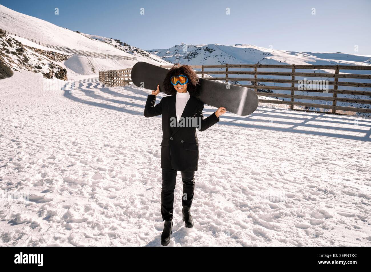 Happy Ethnic Sportswoman en vêtements élégants et lunettes de protection pour le transport du snowboard sur un support enneigé au soleil Banque D'Images