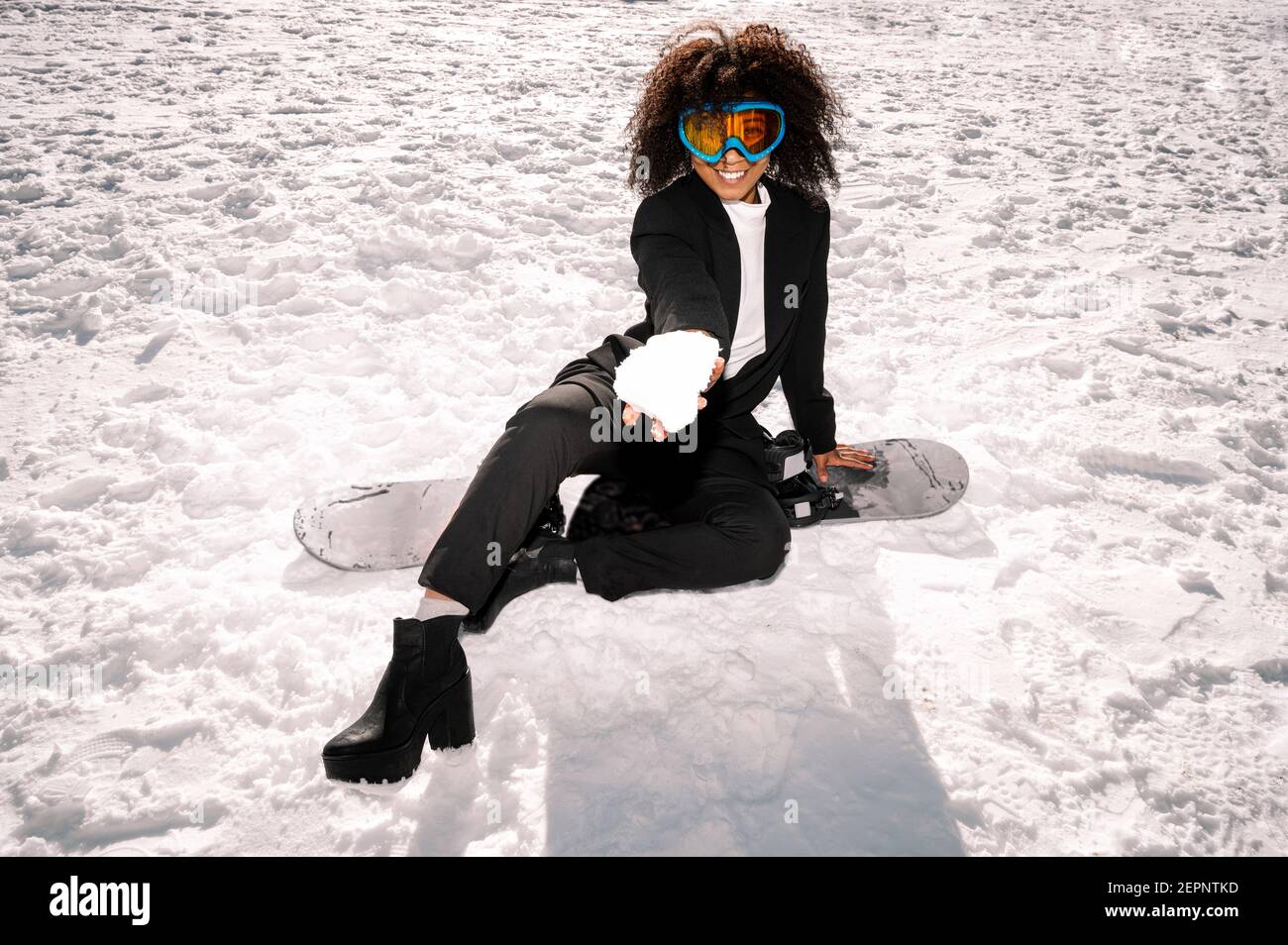 Athlète afro-américain de bonne humeur et de protection lunettes assises sur le snowboard tout en regardant l'appareil photo en hiver Banque D'Images