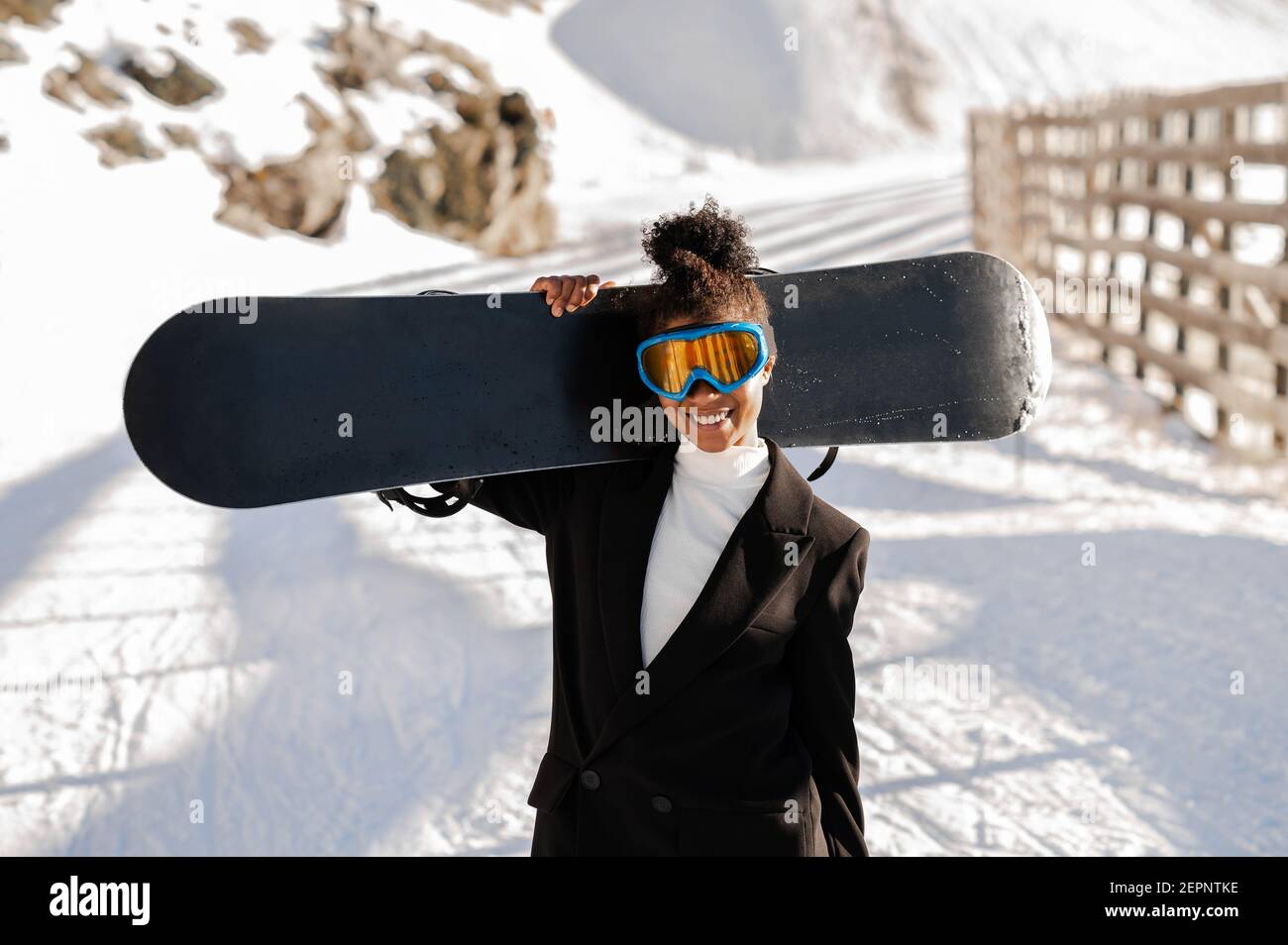 Happy Ethnic Sportswoman en vêtements élégants et lunettes de protection pour le transport du snowboard sur un support enneigé en plein soleil regardant l'appareil photo Banque D'Images