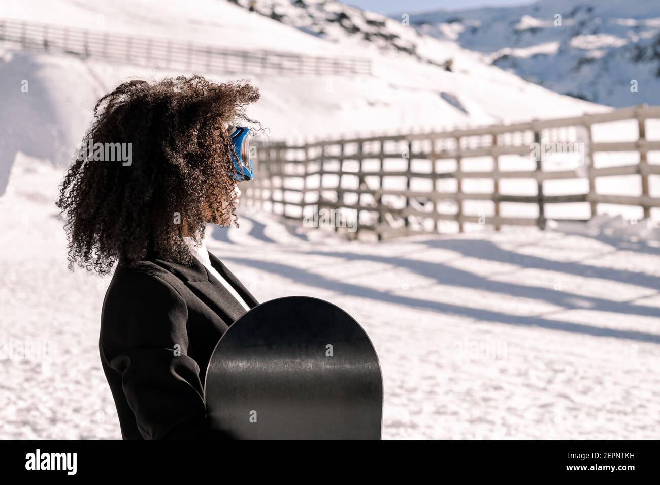 Happy Ethnic Sportswoman en vêtements élégants et lunettes de protection pour le transport du snowboard sur un support enneigé, à la lumière du soleil Banque D'Images