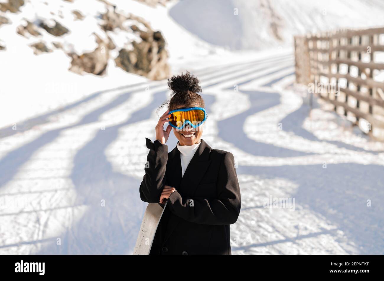 Happy Ethnic Sportswoman en vêtements élégants et lunettes de protection pour le transport du snowboard sur un support enneigé en plein soleil regardant l'appareil photo Banque D'Images