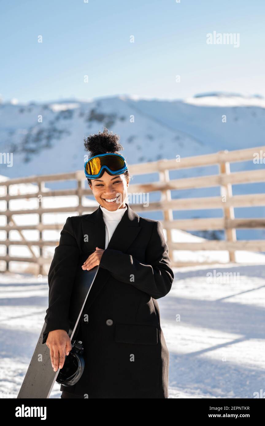 Happy Ethnic Sportswoman en vêtements élégants et lunettes de protection pour le transport du snowboard sur un support enneigé en plein soleil regardant l'appareil photo Banque D'Images