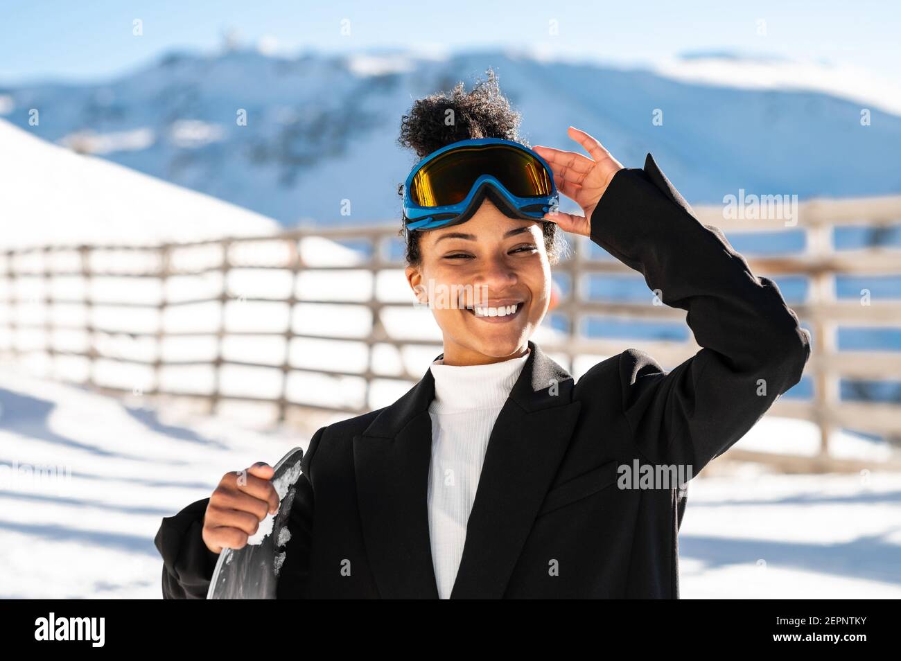 Happy Ethnic Sportswoman en vêtements élégants et lunettes de protection pour le transport du snowboard sur un support enneigé en plein soleil regardant l'appareil photo Banque D'Images