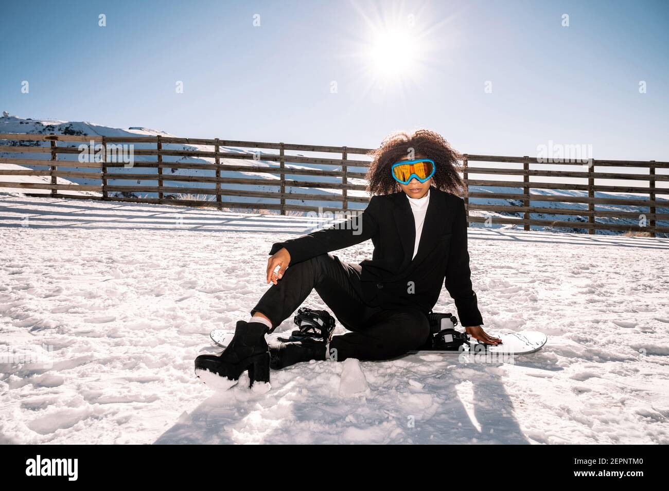 Athlète afro-américain en vêtements à la mode et lunettes de protection assis sur le snowboard tout en regardant l'appareil photo en hiver Banque D'Images
