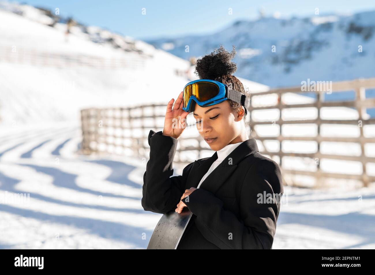 Jolie athlète afro-américaine en tenue tendance et protectrice lunettes debout avec surf des neiges, vue vers le bas en hiver Banque D'Images